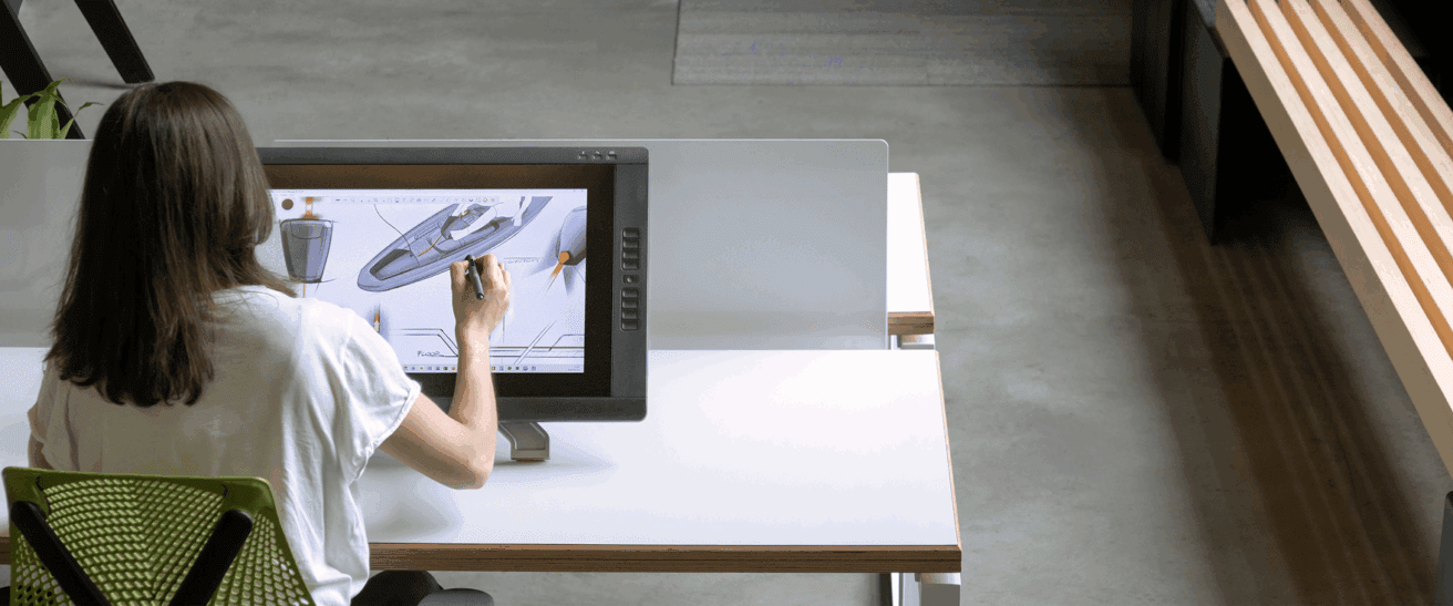 A person with long hair sits at a desk, using a stylus to draw a detailed boat design on a large digital touchscreen tablet in a modern, open workspace.