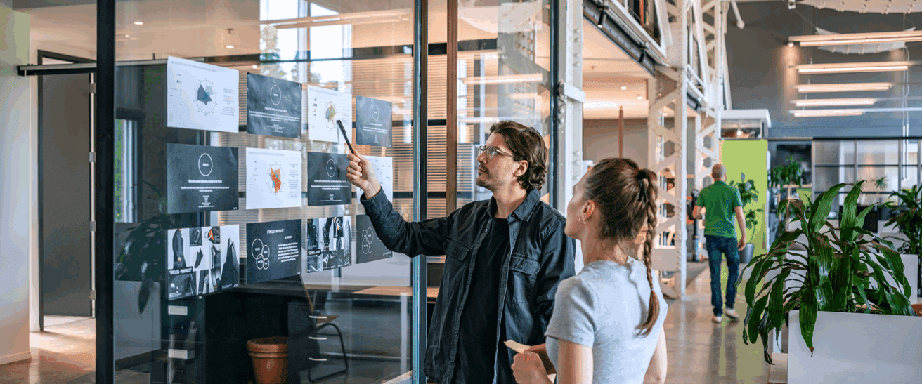 Two people stand in a modern office, looking at graphs and charts posted on a glass wall. One person points to the papers while the other observes, both engaged in discussion.