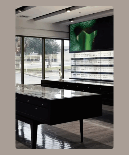 Intérieur d'un magasin de lunettes moderne avec des vitrines, des étagères garnies de lunettes, de grandes fenêtres laissant entrer la lumière naturelle et un écran numérique sur le mur affichant une image verte abstraite.