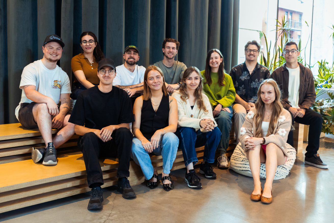Un groupe de onze personnes est assis et debout sur des bancs en bois à l'intérieur, souriant à l'appareil photo. De grandes fenêtres et des plantes vertes sont visibles dans cet espace moderne et lumineux.
