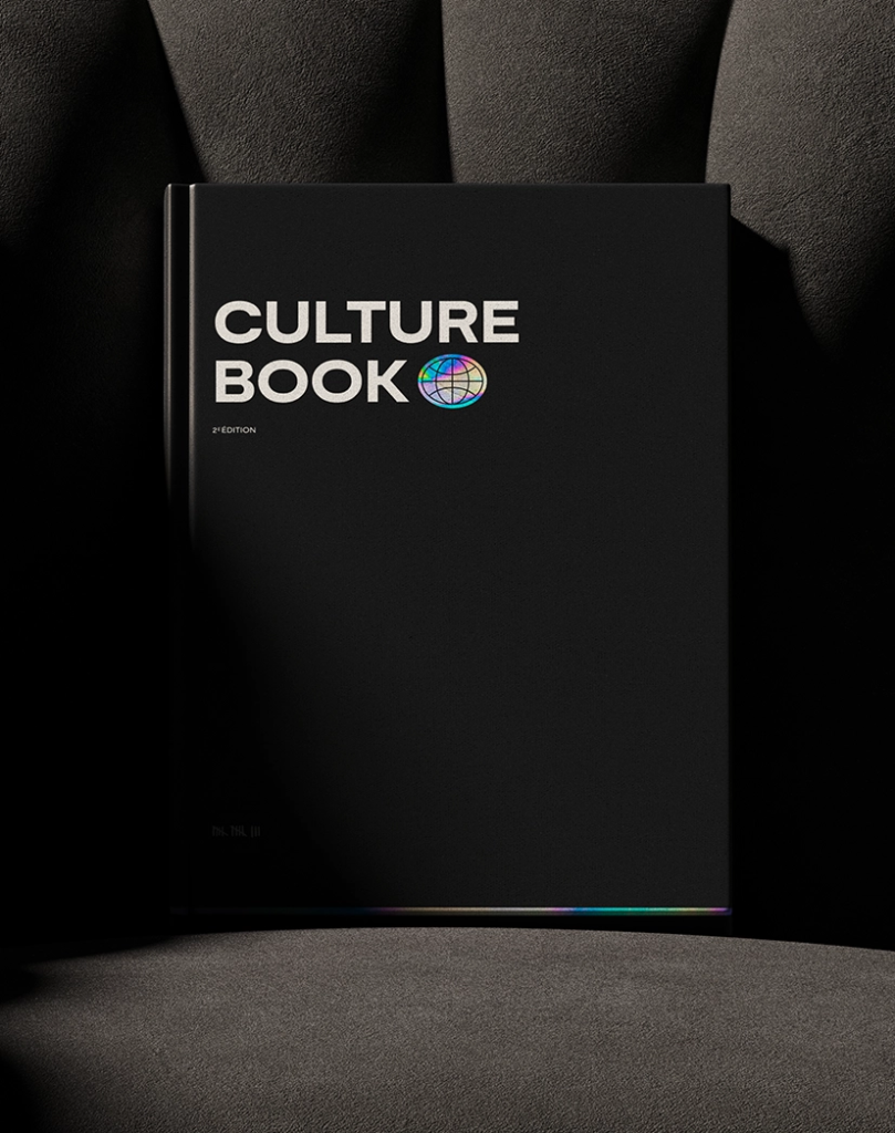 Un livre noir intitulé CULTURE BOOK avec une icône de globe coloré sur la couverture repose sur une chaise sombre et texturée. L'éclairage crée des ombres dramatiques sur le livre et la chaise.