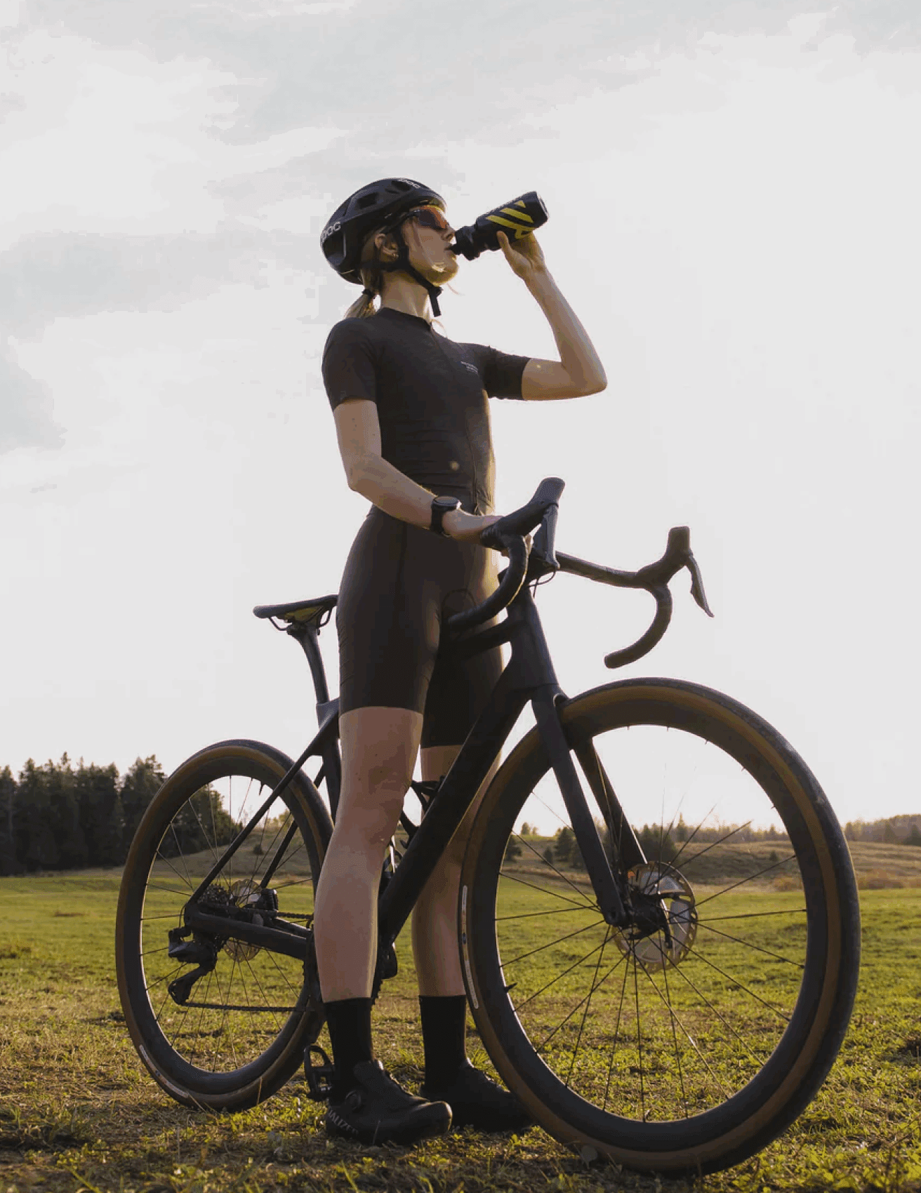 Un cycliste portant un casque et des lunettes de soleil se tient à l'extérieur avec son vélo, buvant une bouteille d'eau. L'arrière-plan montre un champ ouvert et herbeux et une ligne d'arbres lointaine sous un ciel lumineux.