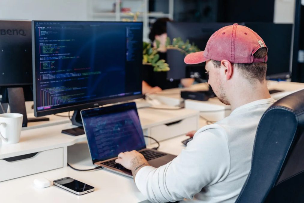 Un homme portant une casquette rouge travaille à un bureau équipé d'un ordinateur portable et d'un grand écran affichant du code. Un téléphone, une tasse et des écouteurs sans fil se trouvent sur le bureau. Une autre personne apparaît floue à l'arrière-plan.